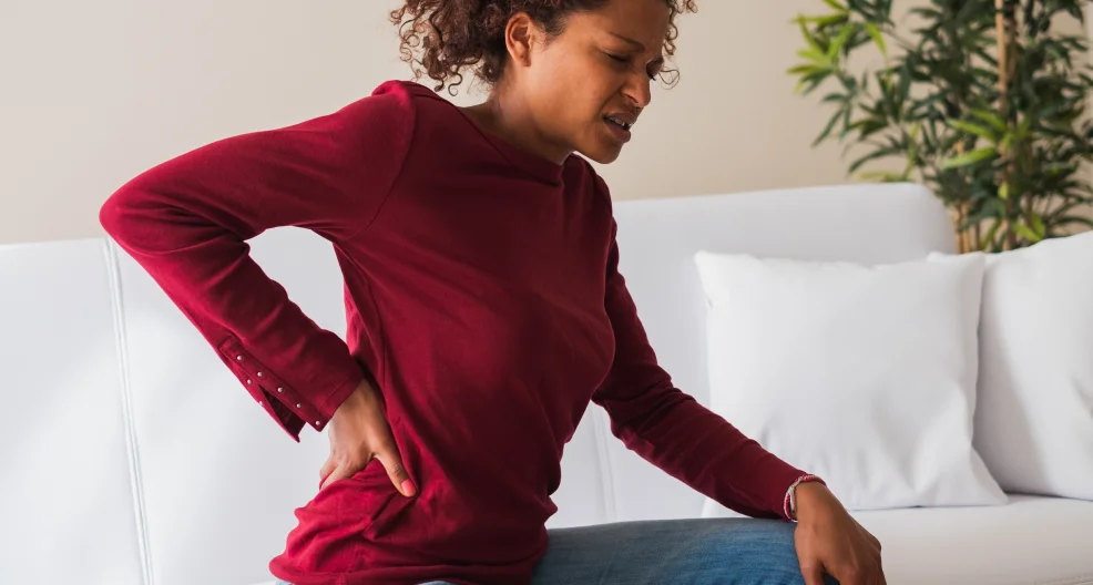 Young woman sitting in her couch, suffering from a back pain cramp.